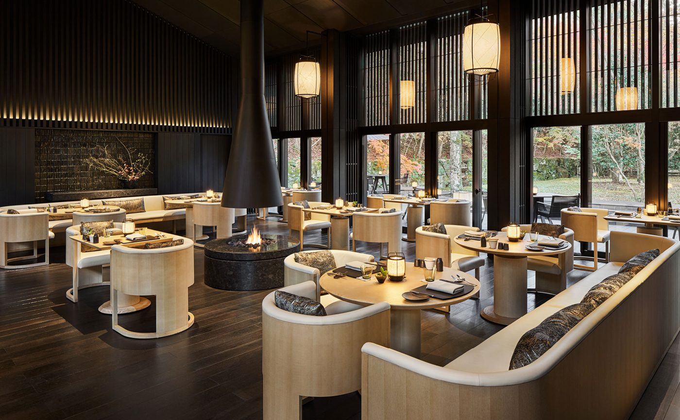 Interior of The Living Pavilion by Aman at Aman Kyoto, with a black conical hearth in the centre, pale timber tables, paper lanterns and floor-to-ceiling windows onto the forest, courtesy of Aman