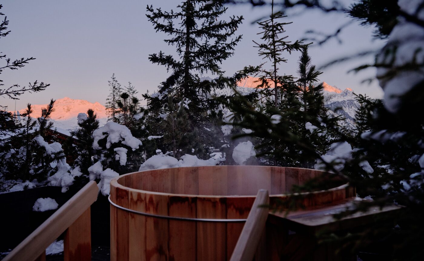 A wooden hot tub on a suite terrace at Aman Le M&eacute;l&eacute;zin at dusk, above snow-covered pine forest
