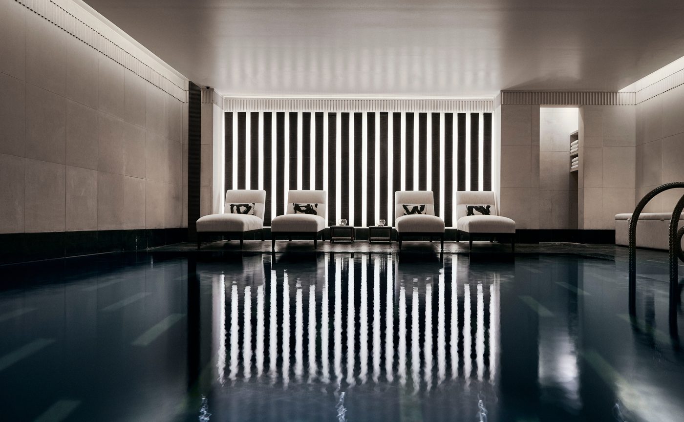 The indoor pool at Aman Spa at The Connaught in Mayfair, four pale loungers reflected in dark water beneath a wall of vertical light, courtesy of Aman