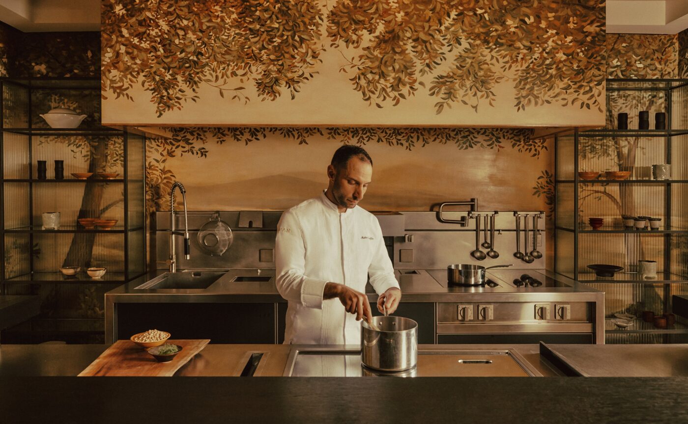 Executive Chef Matteo Panfilio alone at the stove of The Palazzo Kitchen Table, the painted foliage of the room rising behind the steel kitchen, courtesy of Aman