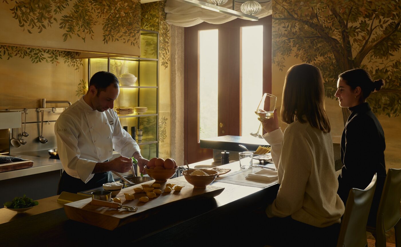 Executive Chef Matteo Panfilio at the counter of The Palazzo Kitchen Table at Aman Venice, plating a first course for two guests beneath walls painted with trees, courtesy of Aman
