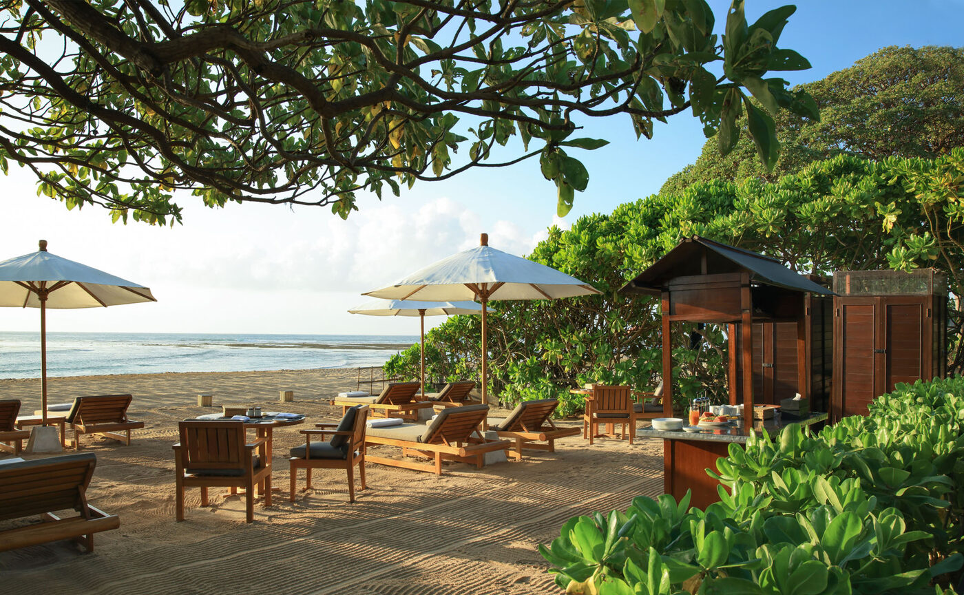 The Beach Terrace at Aman Villas at Nusa Dua at first light, teak loungers and white parasols on the sand beneath sea almond trees, courtesy of Aman