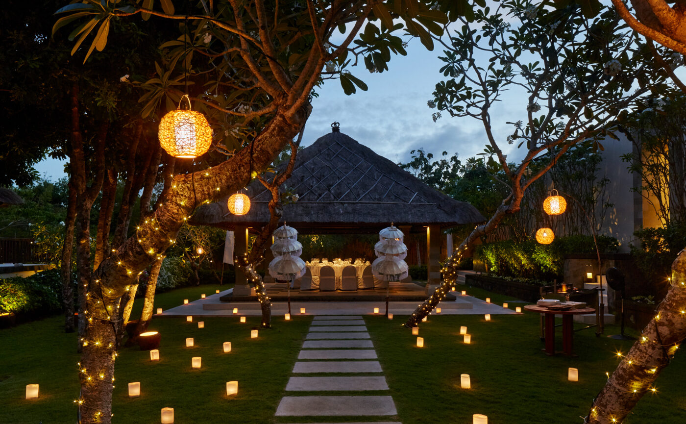 A candlelit dining bale at dusk in the garden of Aman Villas at Nusa Dua, lanterns hung in the frangipani trees and paper lamps along the lawn, courtesy of Aman