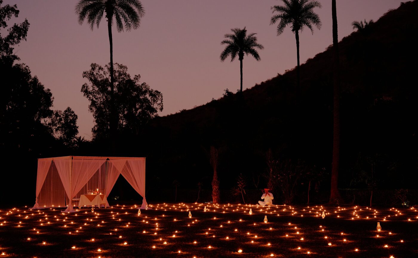 A candlelit dinner table laid on the lawns at Amanbagh at dusk, palm trees silhouetted against the mauve Aravalli Hills, courtesy of Aman