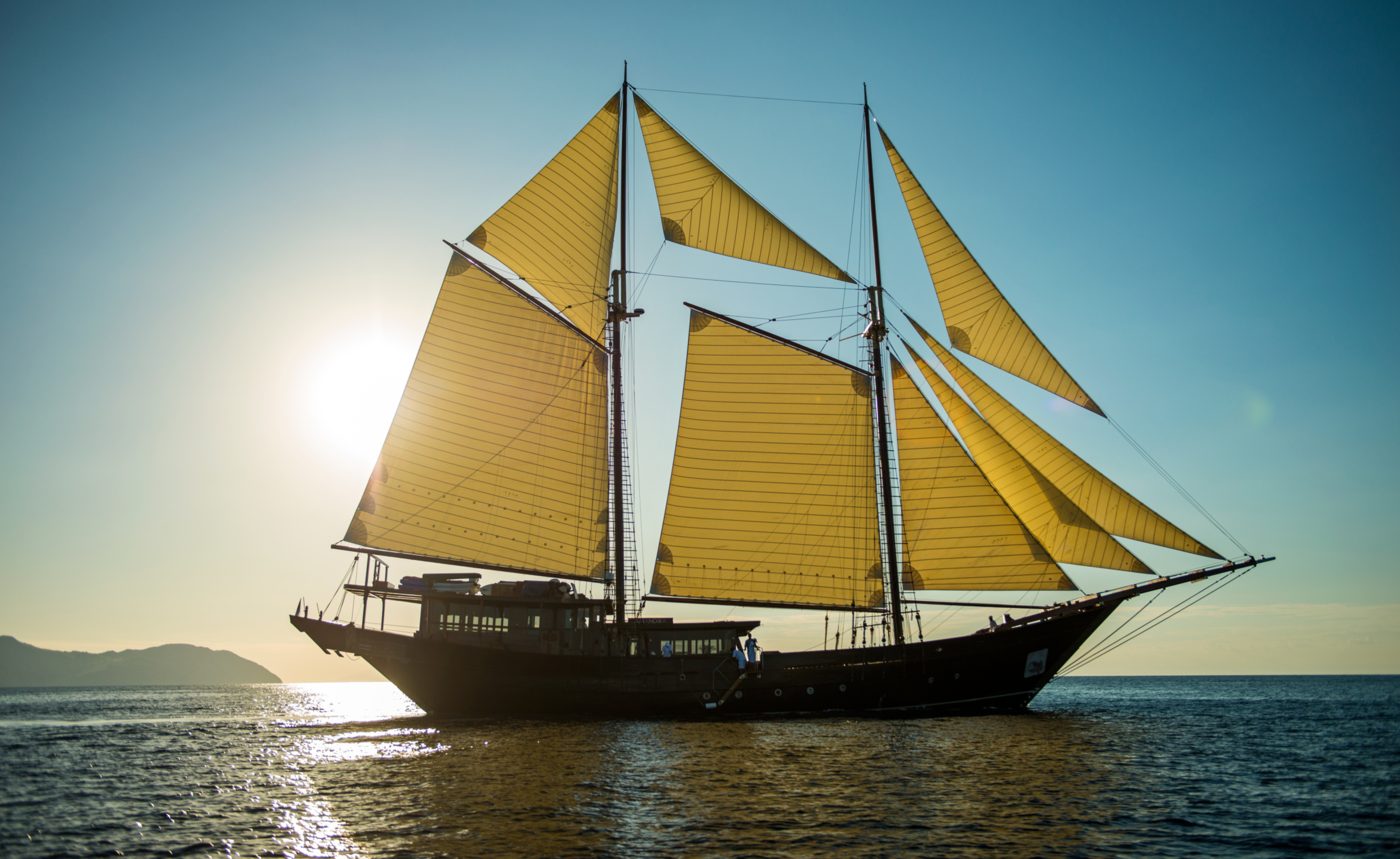 Amandira under sail at dusk in the East Indonesian archipelago, a two-masted wooden Pinisi with golden sails raised against a pale evening sky, courtesy of Aman