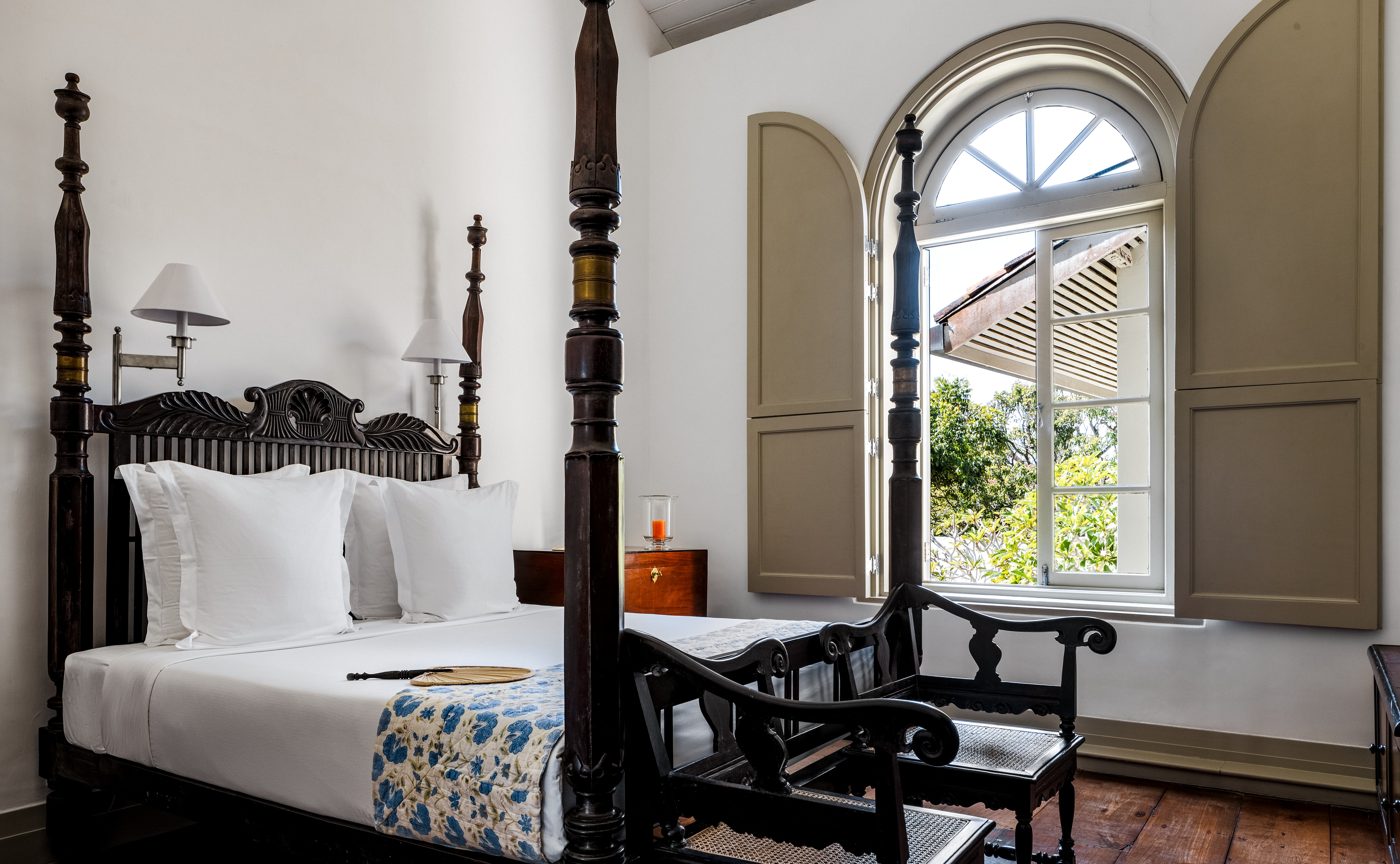 A guest room at Amangalla with a dark carved four-poster bed, white linen, antique colonial chairs and tall arched windows with painted shutters open to the trees, courtesy of Aman