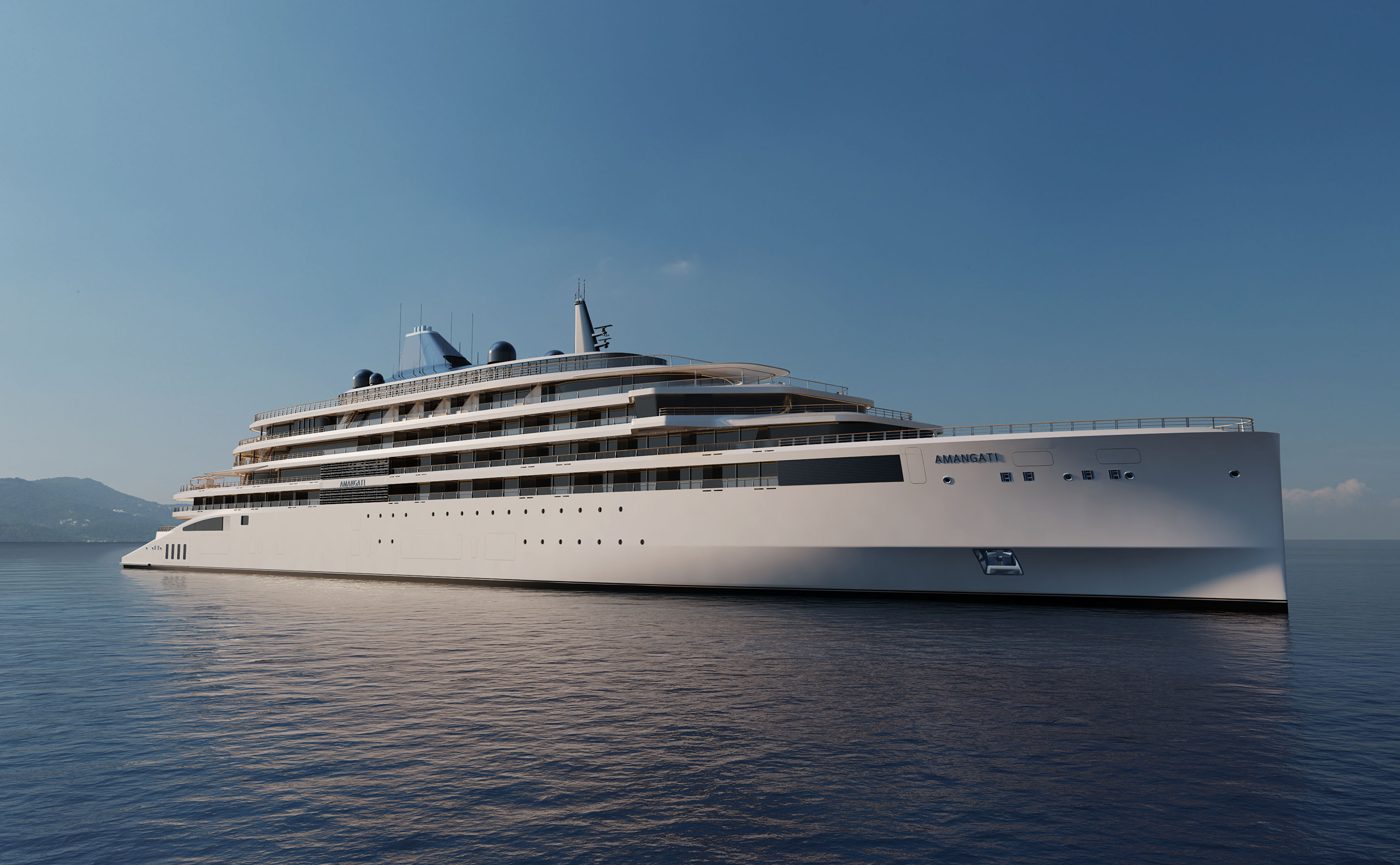 Amangati, the first yacht from Aman at Sea, a white nine-deck motor yacht at anchor on a calm sea with a mountainous coast behind, courtesy of Aman at Sea
