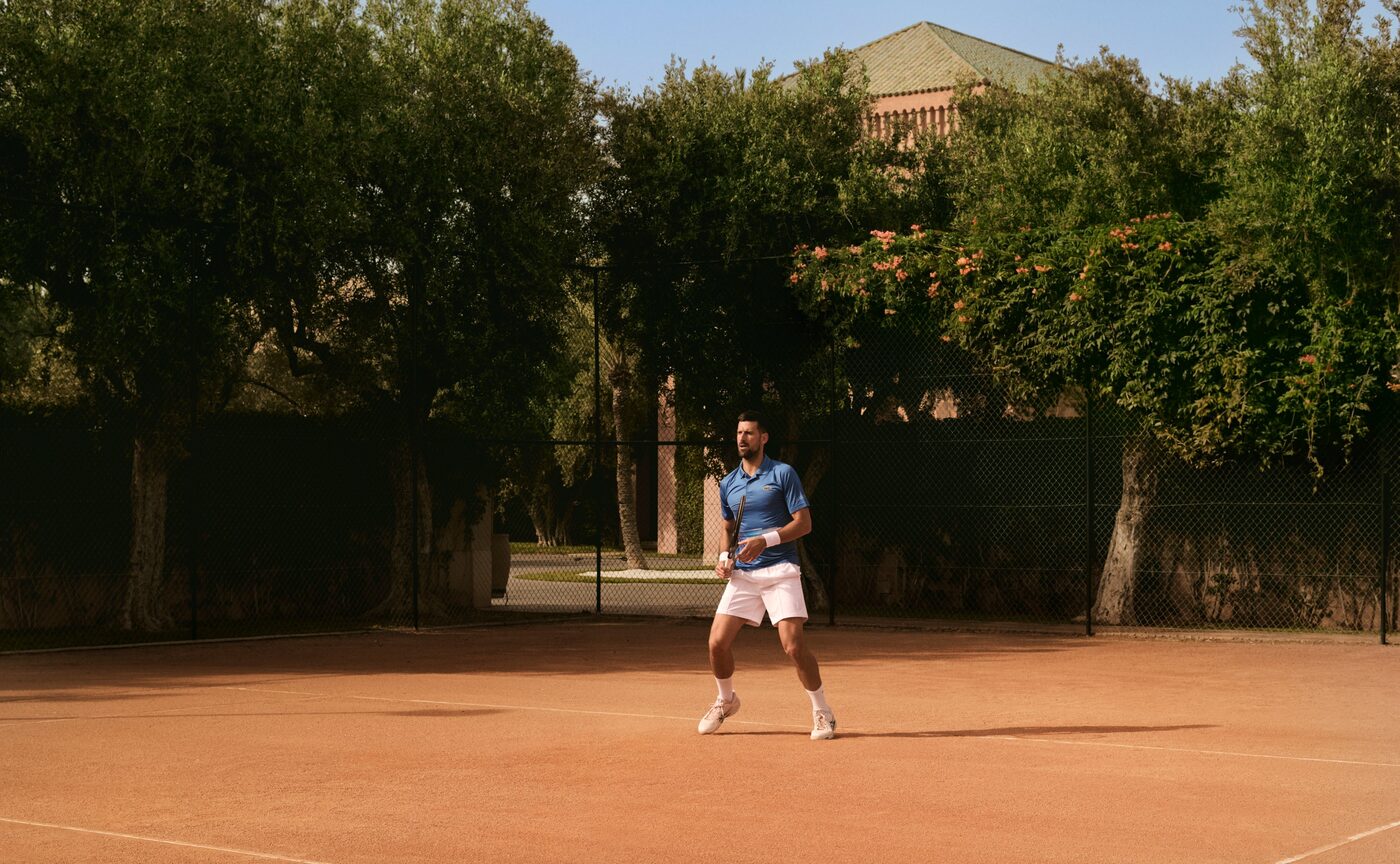 Novak Djokovic on the clay tennis court at Amanjena in Morocco, framed by green vegetation, courtesy of Aman
