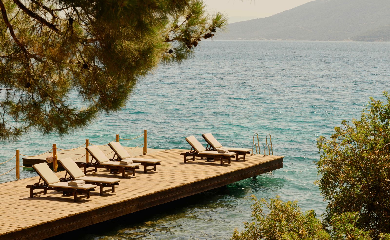 Empty teak loungers on the wooden jetty at Amanruya, Bodrum, under pine branches above the turquoise Aegean, courtesy of Aman