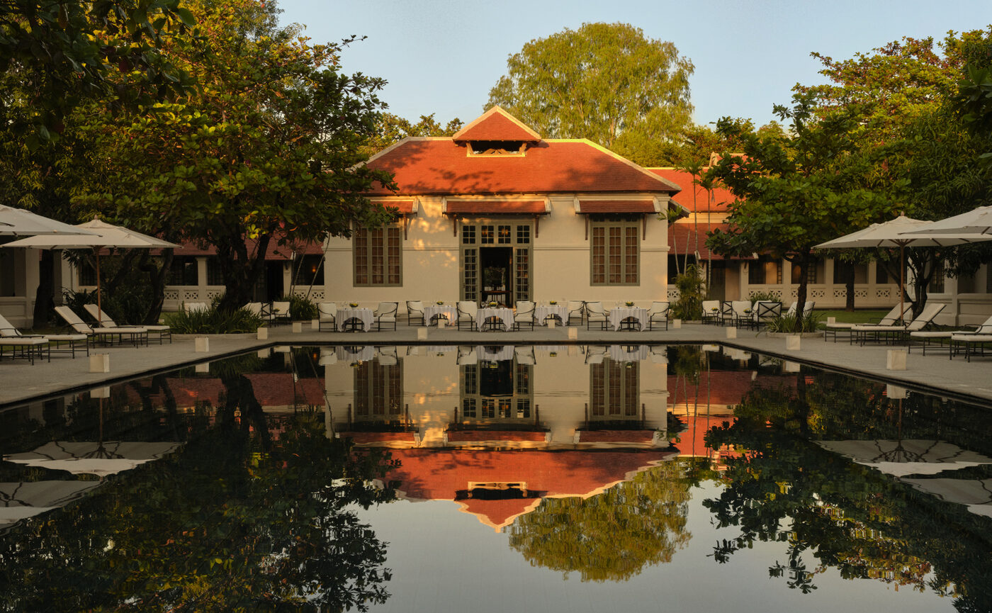 A dinner table set at the edge of Amantaka's main pool at dusk, the colonial pavilions reflected in the water, courtesy of Aman