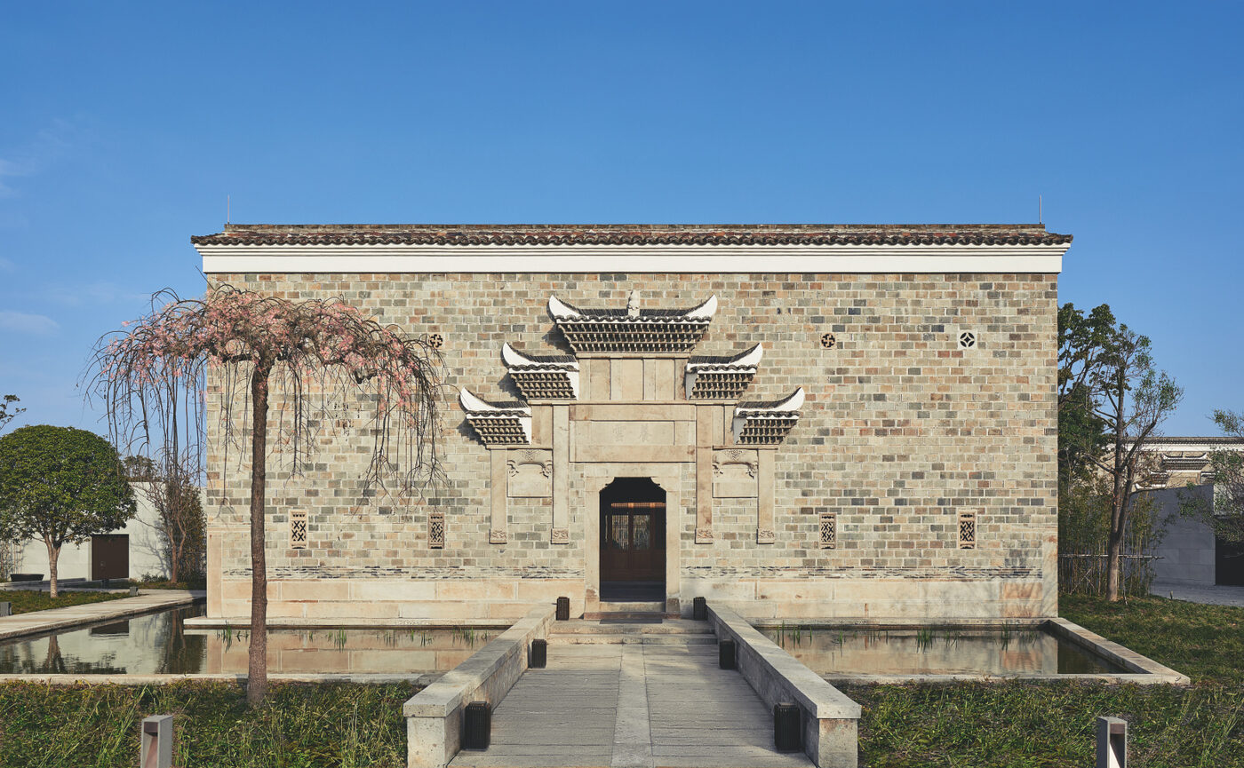A restored Ming-era brick villa at Amanyangyun, its carved stone gateway and upturned eaves mirrored in a still reflecting pool, a weeping blossom tree to one side, under a clear Shanghai sky
