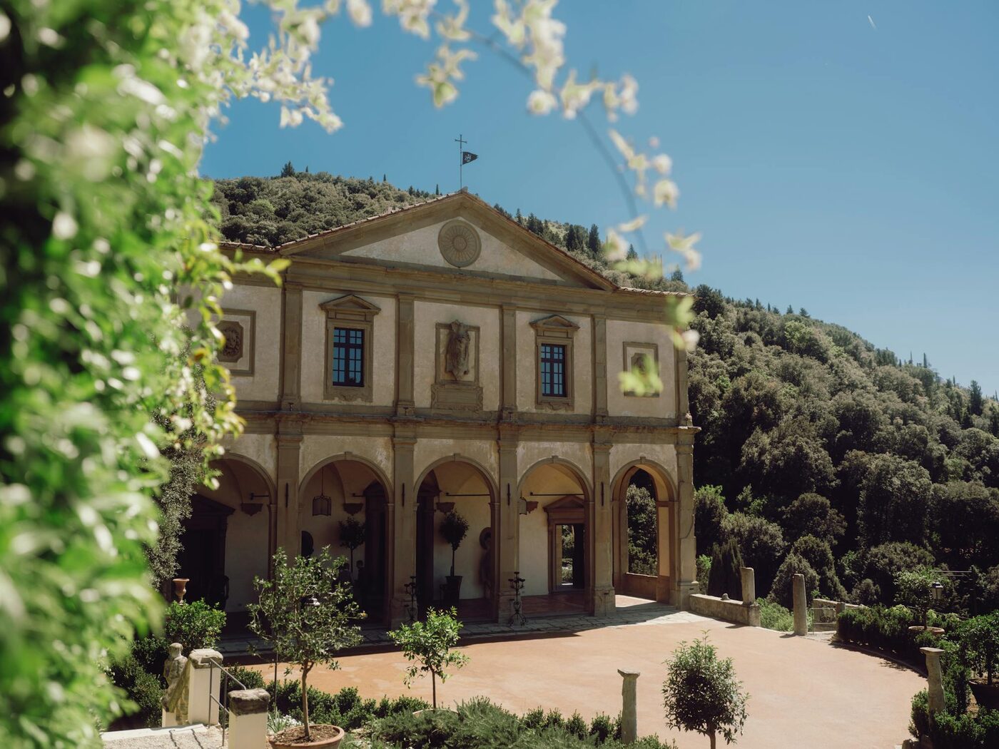 The stone facade and arched loggia of Villa San Michele in Fiesole, seen through white blossom, with the wooded hillside of Monte Ceceri behind, courtesy of Belmond