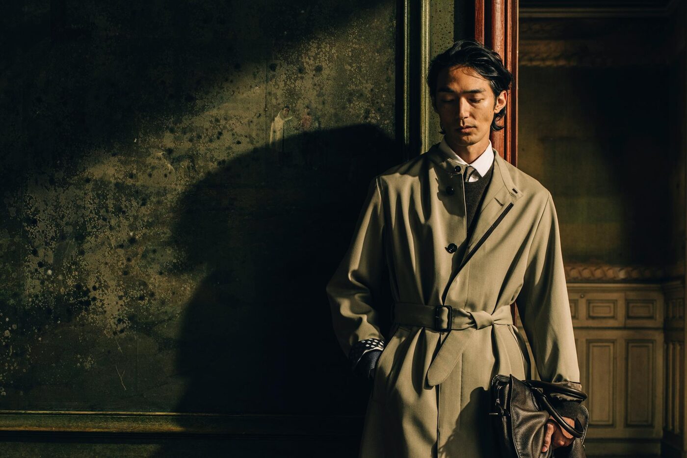 A model in a belted stone-coloured Berluti coat holding a leather bag, standing against a darkened fresco wall at the Fondation Simone et Cino Del Duca in Paris