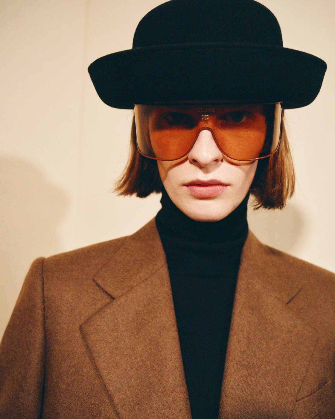 A Celine Automne 2026 look by Michael Rider: camel coat, black bowler hat and amber Celine shield sunglasses