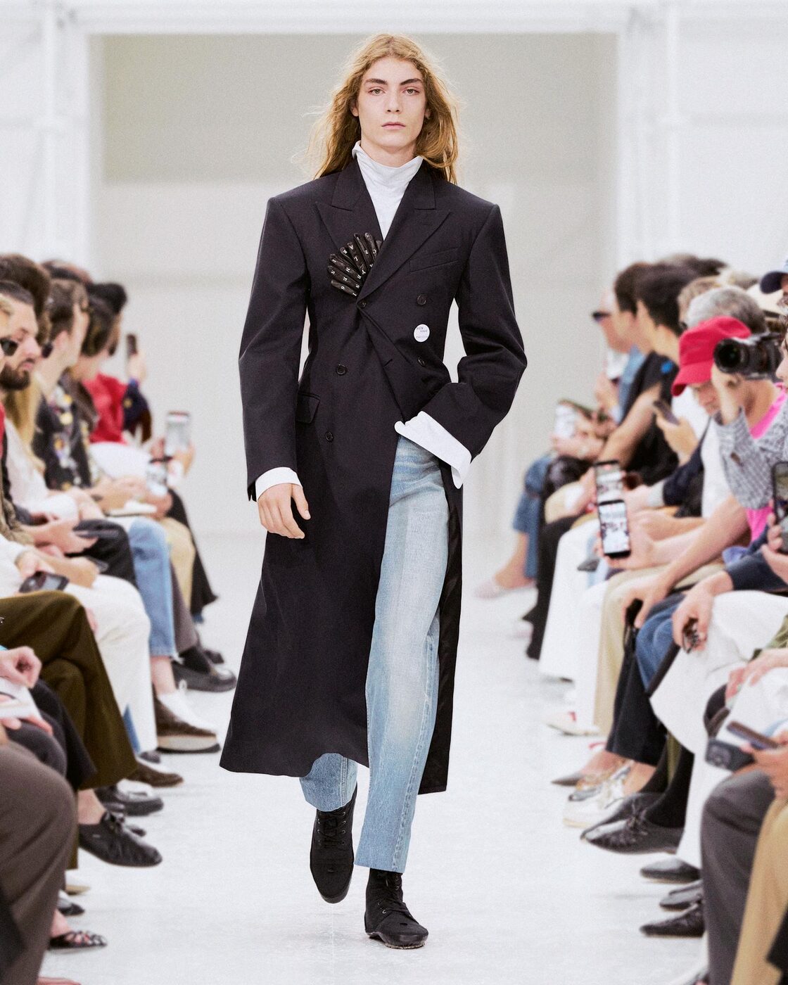 A model in a long navy double-breasted coat over faded jeans, with leather gloves fanned out of the chest pocket, at the Celine men's spring 2027 show