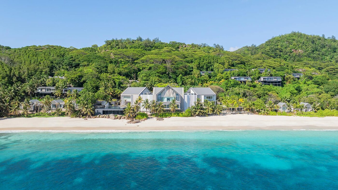 Aerial view of Cheval Blanc Seychelles, its gabled pale-timber villas set into the jungle hillside above the white sand and turquoise water of Anse Intendance on Mahé