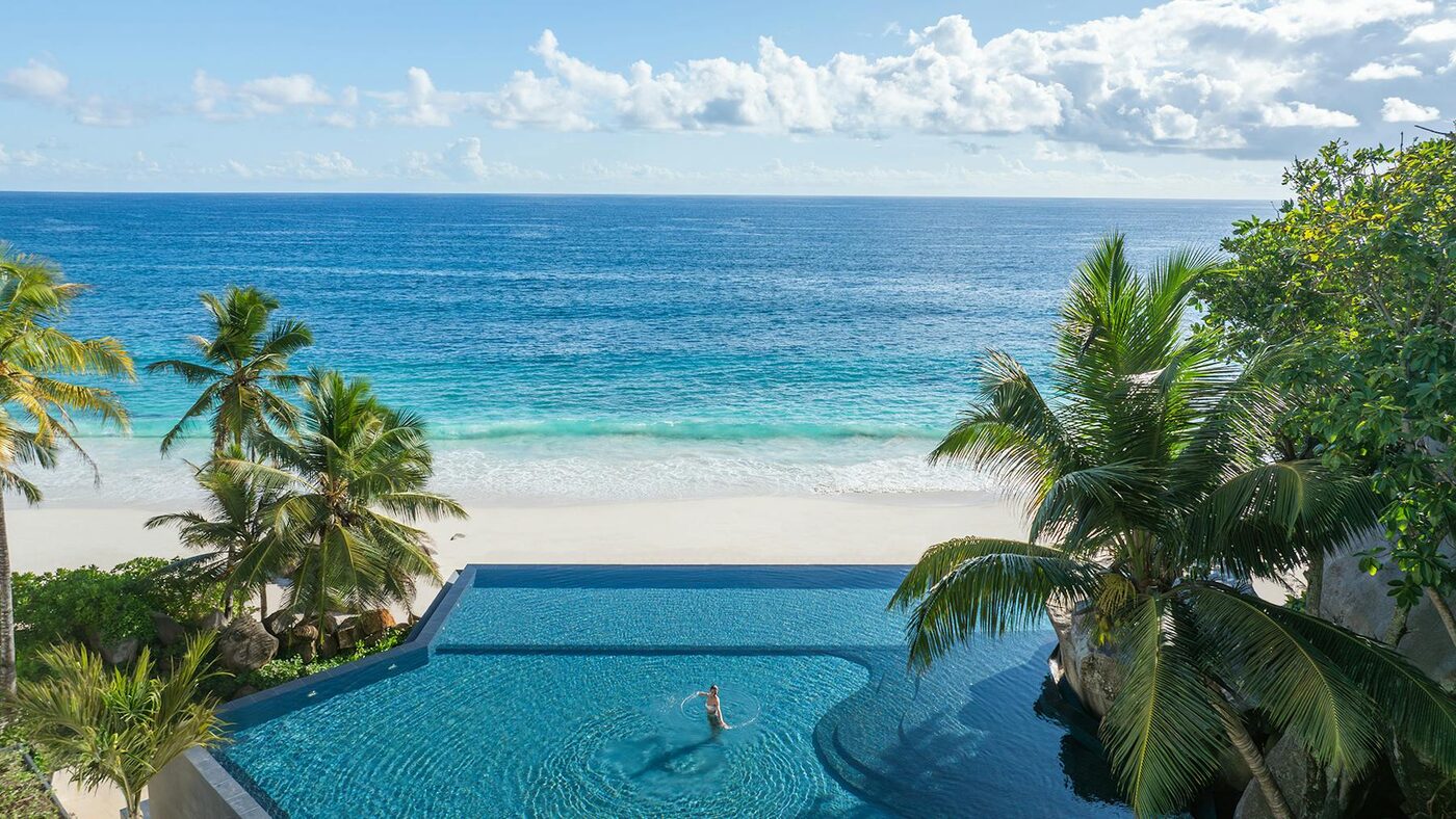 A two-tier infinity pool at Cheval Blanc Seychelles spilling toward Anse Intendance beach and the Indian Ocean, framed by palms and granite boulders