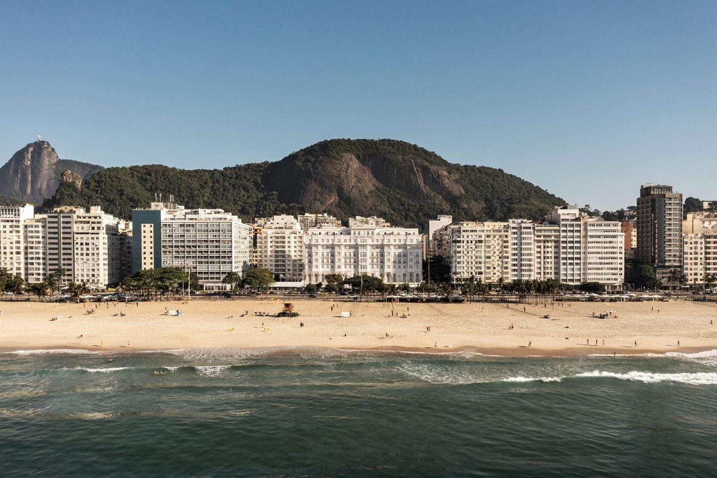 The Copacabana Palace seen from the water, its cream facade set among the apartment blocks of Avenida Atlantica with Copacabana beach in front and the forested hills of Rio behind, courtesy of Belmond