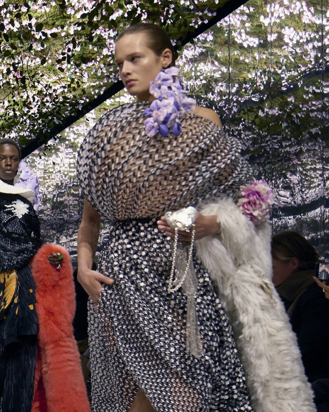 A Dior Fall Winter 2026 couture look in pleated metallic tweed with a violet floral corsage and a pearl-strung bag