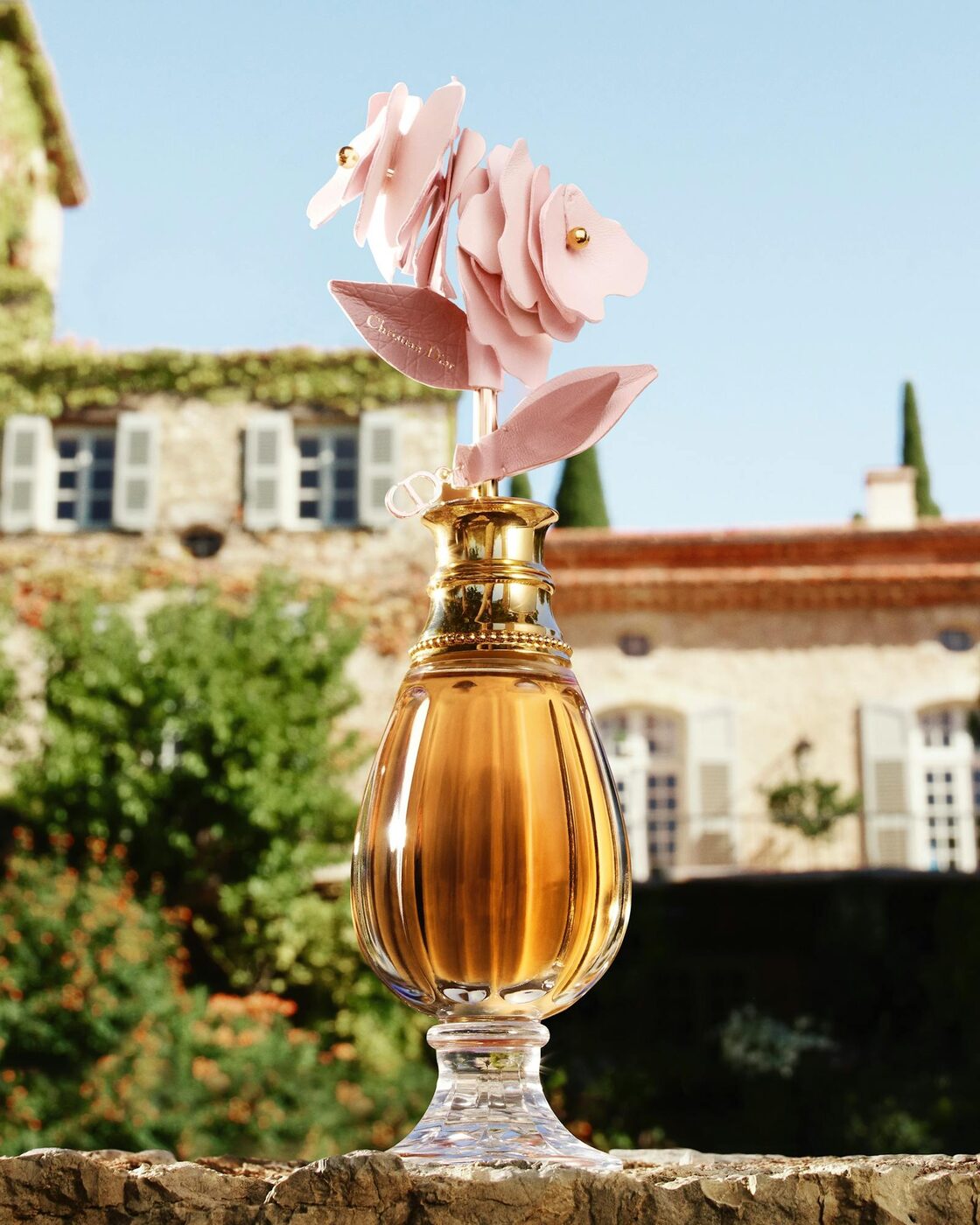 A fluted amber Dior flacon crowned with a pale pink rose crafted from layered lambskin leather, a leaf embossed with the cannage motif and the Christian Dior signature, set against a Provençal château garden