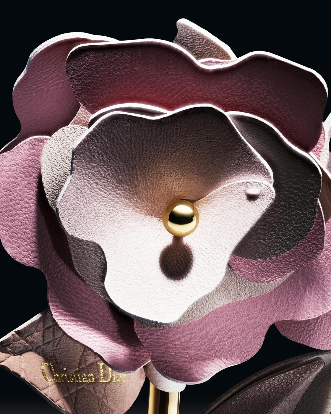 Close view of the Dior leather rose, its lambskin petals graded from wine to powder pink around a gilded bead stamen, with the Christian Dior name embossed in gold on a cannage leather leaf