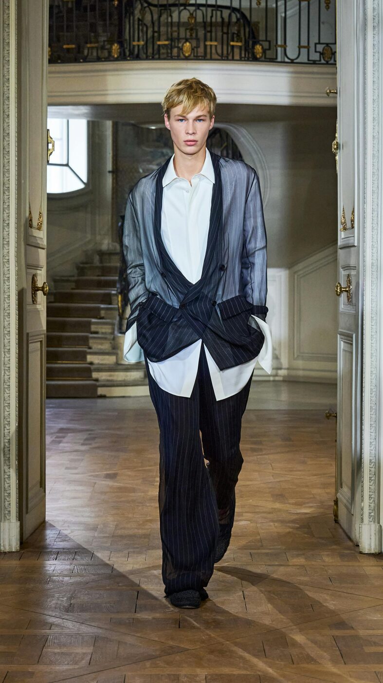 A model in a translucent pinstripe Dior jacket printed on silk chiffon, worn over a white shirt and pinstripe trousers, walking through the panelled interior of the Musée Nissim de Camondo