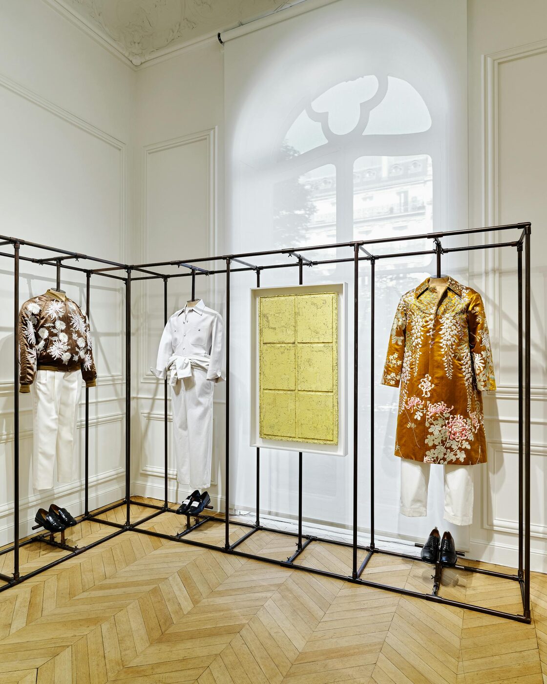 Givenchy Spring-Summer 2027 menswear on black metal racks inside the Avenue George V salons: an embroidered bomber, a white workwear set and a floral evening coat beside a gold Rachel Whiteread cast