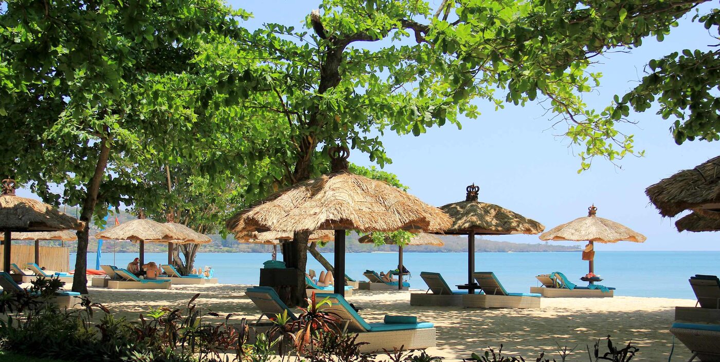 Thatched sun umbrellas and loungers under trees on the sand at Jimbaran Bay, Bali