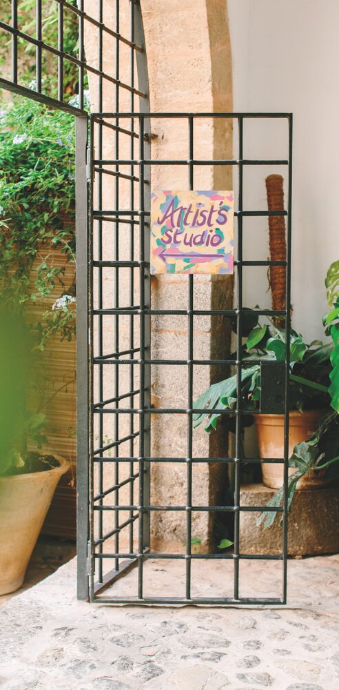 The wrought-iron gate to the Artist's Studio at La Residencia, Deià, a hand-painted sign fixed to the grille beside potted plants and a stone archway