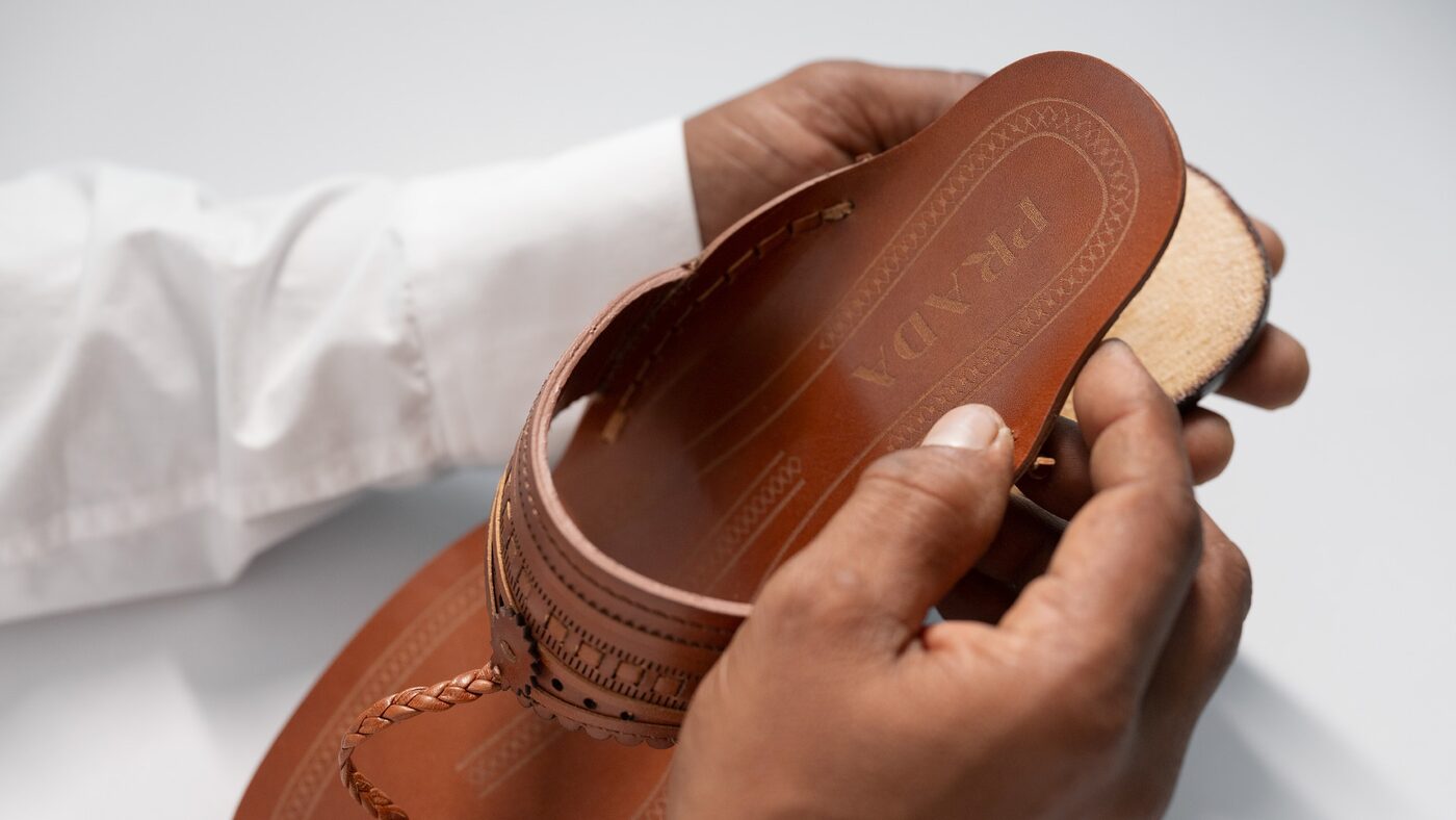 Hands holding a tan leather Kolhapuri-style sandal with the Prada name embossed on the insole