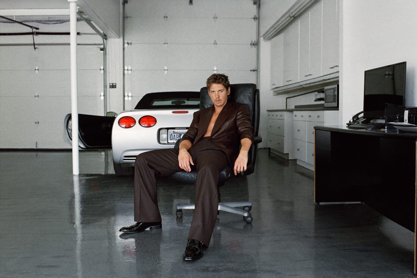 A Saint Laurent Fall 2026 menswear campaign image, a man in a brown leather blazer and pinstripe trousers seated in a black chair in a bare white garage with a white convertible behind him