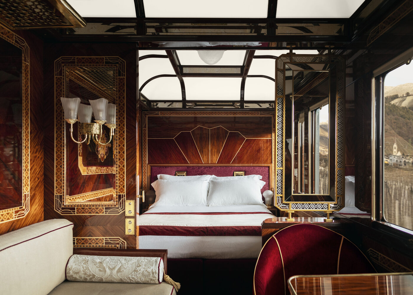 A suite aboard the Venice Simplon-Orient-Express, with fan-patterned marquetry panelling, a brass wall light, mirrored doors and a made bed beside the window, courtesy of Belmond